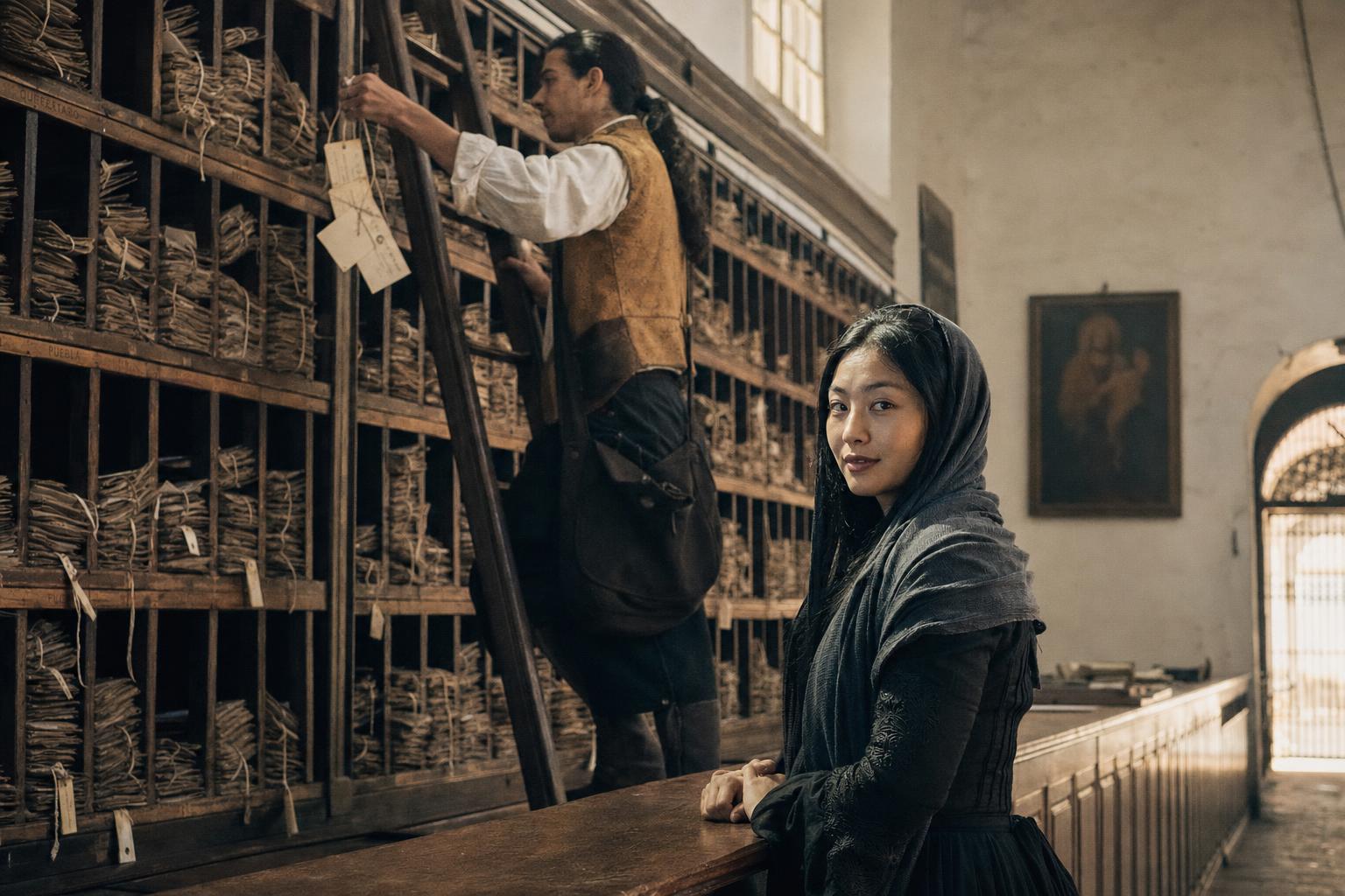 Woman in dark shawl stands at a wooden counter in an old archive room while a man climbs a ladder to reach shelved documents.